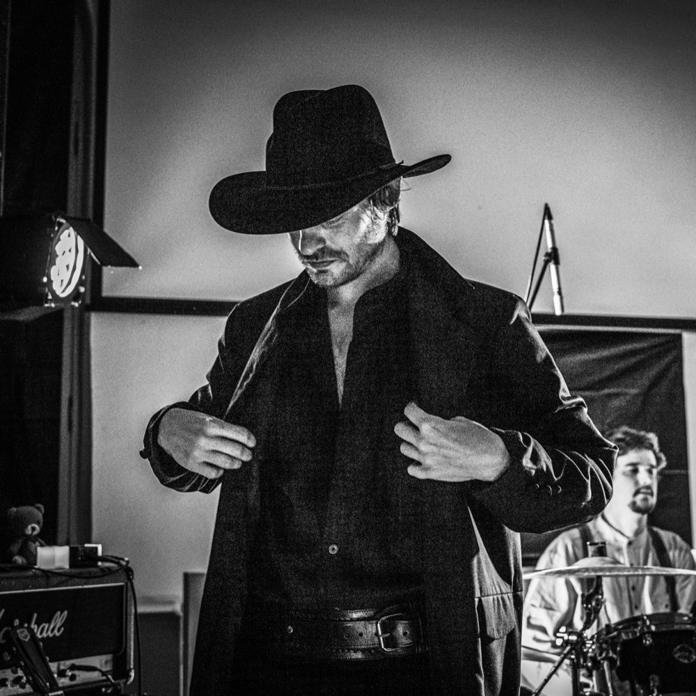 Chris Green in a black coat and hat, adjusting his collar on stage